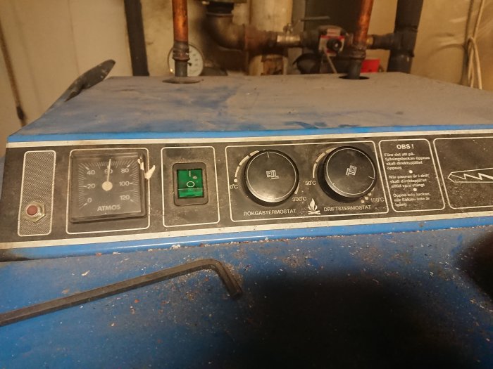 Kontrollpanel på värmepanna med termometer, strömbrytare och termostater mot suddig bakgrund.