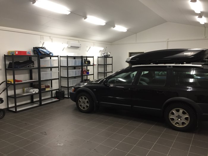 Ett garage med grå plattor på golvet, en parkerad bil och svarta hyllor lastade med diverse föremål.