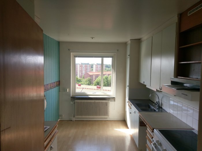 En ljus köksinteriör före renovering med parkettgolv, vita skåp och ett fönster med utsikt över stadsbebyggelse.