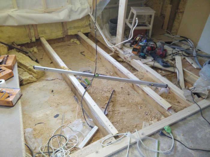 Rakennustyömaa, jossa lattiaremontti käynnissä. Puisia palkkeja asetettu paikalleen, vieressä työkaluja ja sähköjohtoja. Lattiatason mittaus kesken.