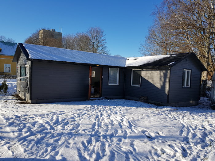 En gråmålad stuga med snötäckt tak och omgivande snö i ett soligt vinterlandskap.