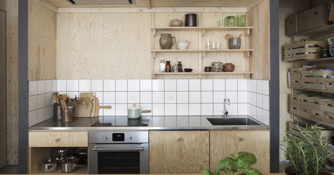 Modernt kök med ljusa plywoodskåp, vit kakelvägg, rostfri bänkskiva och öppna hyllor.