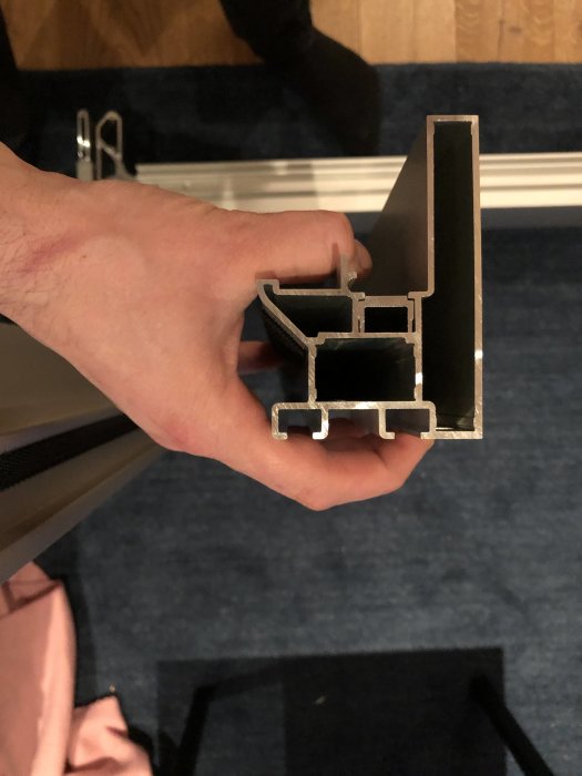 Hand håller ett tvärsnitt av en aluminiumprofil mot en suddig bakgrund.
