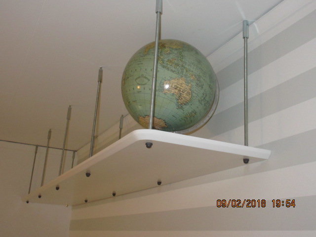 Vit hylla monterad på vägg med skjutbara stänger och en glob på toppen, framför en vit vägg med horisontella linjer.