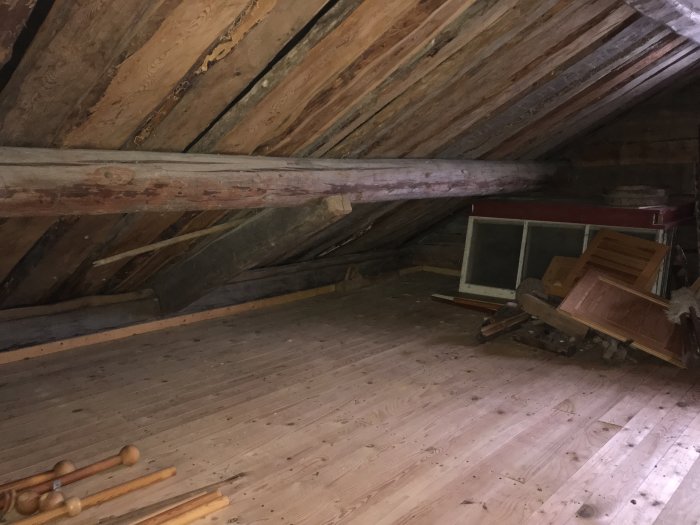 Oinredd vind med synliga takbjälkar, trägolv och gamla fönster samt byggmaterial på ett hus från 1860.