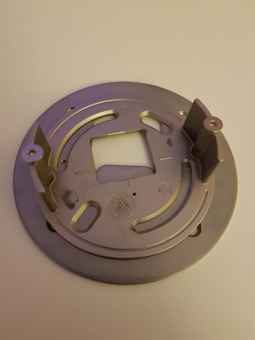 Metallplatta med utstansade hål, fästelement och en krok, tänkt för elektriskt drivdons montering.