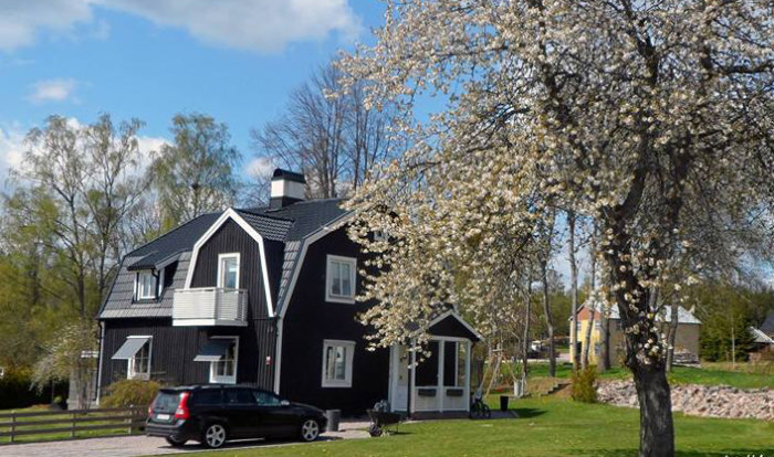 Svartmonterat hus med vita fönsterkarmar och svart bil framför, bredvid blommande träd i soligt väder.