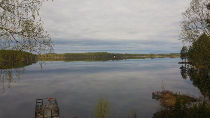 Vy över en stilla sjö i Gästrikland med spegling av skog och himmel i vattnet, flotte i förgrunden.