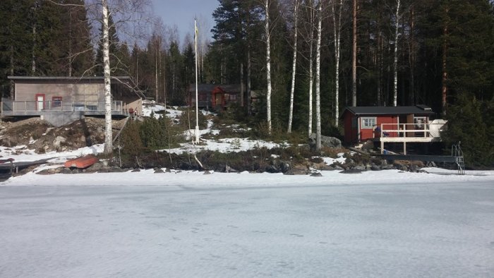 Timmerstuga och modern stuga vid snötäckt sjö i Gästrikland, med solfångare och omgivande skog.