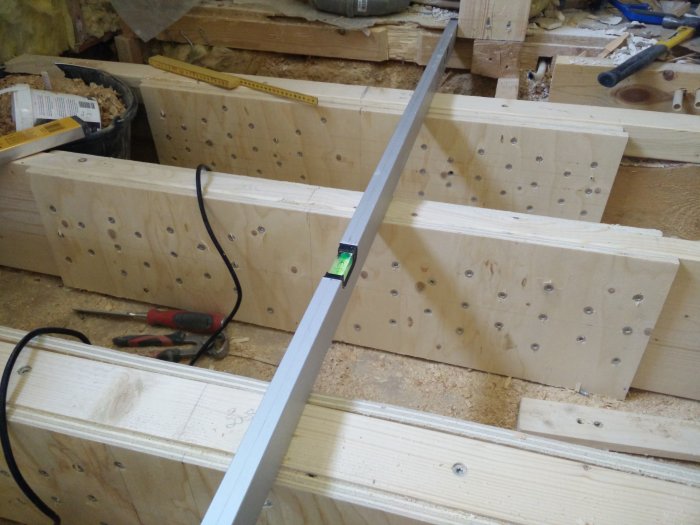 Puupalkit vaakatasossa, poraustyökaluja ja vesivaaka näkyvät. Rakennusprojekti meneillään, riittävä tila alustan valmisteluun.