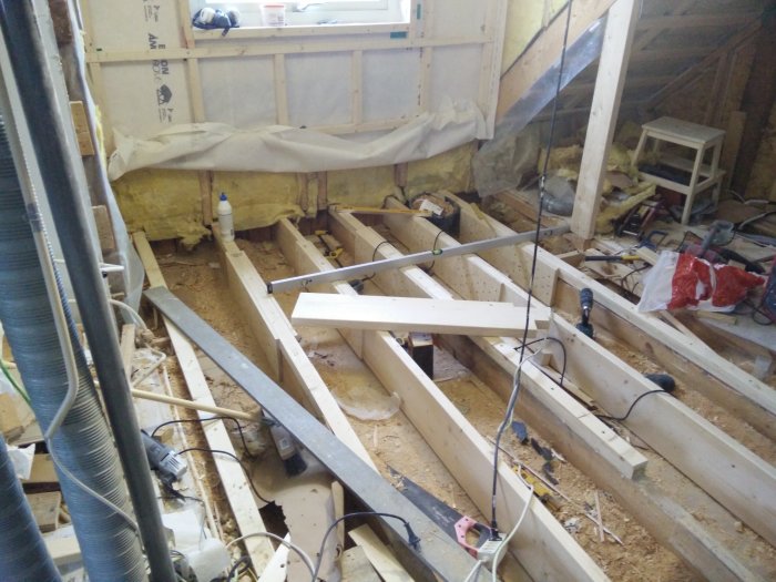 Rakennustyömaa, jossa näkyy puupalkkeja lattiatasossa, poria ja työkaluja lattialla, seinässä eristeitä ja ikkunan alla tarvikkeita.