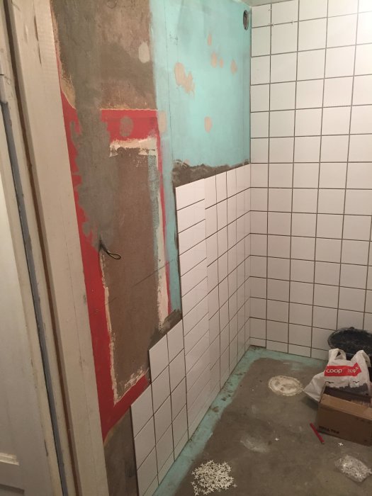 Badrum under renovering med halvsatt vitt kakel, betongväggar och synlig golvslinga.