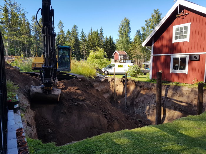 Grävmaskin gräver stort hål för vinkällare vid röd stuga, med grannhus i bakgrunden.