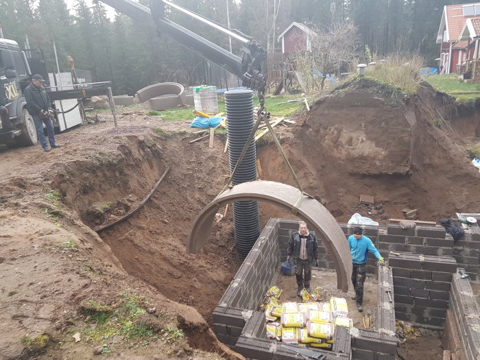 Arbetsplats för bygge av vinkällare med arbetare, leca-block, isoleringsmaterial och grävmaskin.