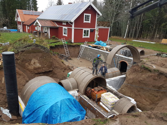 Byggplats med betongrör för vinkällare under konstruktion, arbetsmän och utrustning syns.