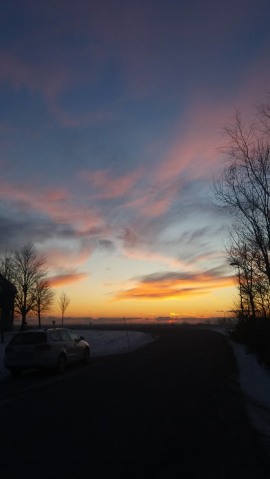 En bil parkerad vid en väg med en dramatisk solnedgång och färggranna moln på vinterhimlen.