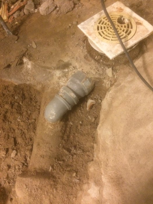 Avloppsrör i gjutjärn, plast och betong delvis grävda i jord med golvbrunn i bakgrunden.