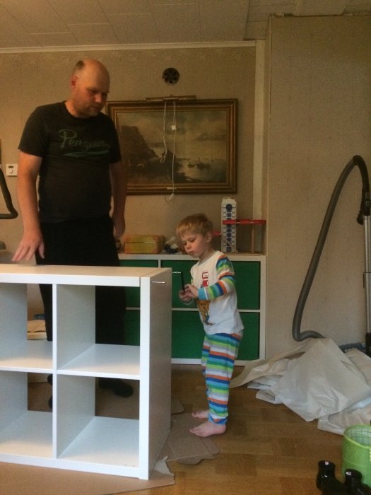 Man och barn i pyjamas monterar en vit förvaringshylla i ett rörigt rum.