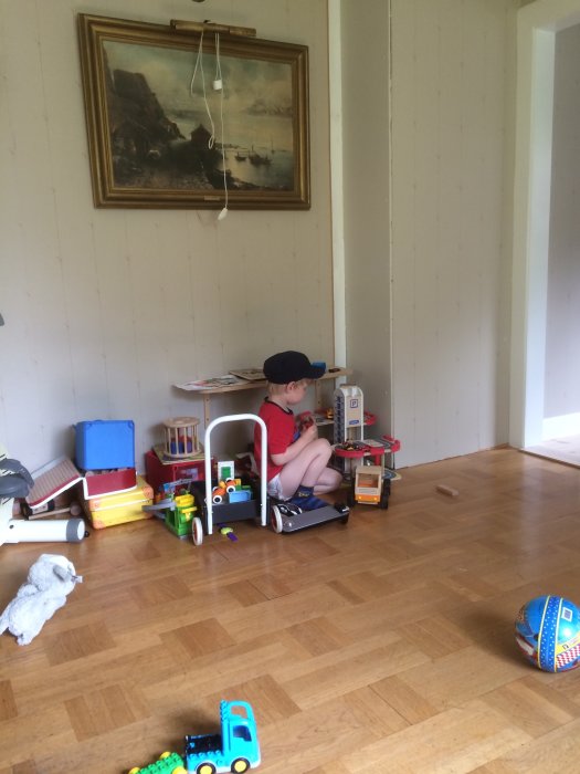 Barn leker på ett trägolv i ett rum med ljus väggpanel och en tavla, platsen där kaminen ska installeras.