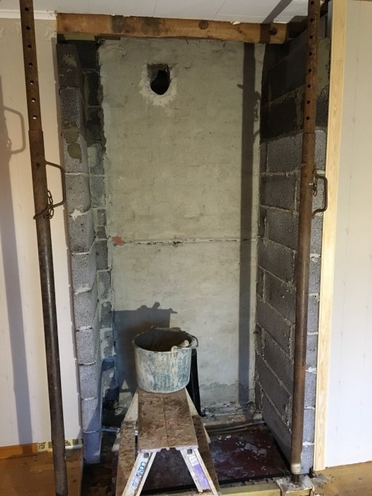 En vägg förberedd för installation av kamin, med stagade väggar, en påbörjad mur samt en stege och hink framför.