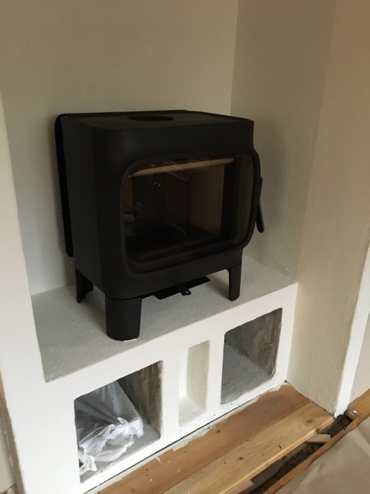 Svart Jøtul F305 kamin installerad på en vit putsad mur med utskurna fack för vedförvaring.