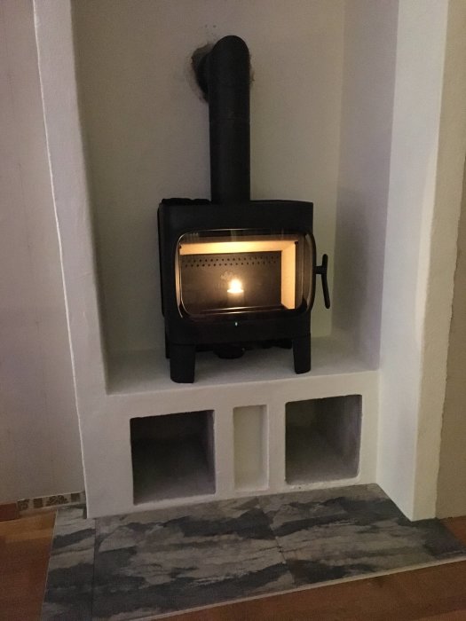 Jøtul F305 kamin med korta ben monterad i ett hörn på en upphöjd putsad bänk med utrymme för ved och klinkergolv framför.