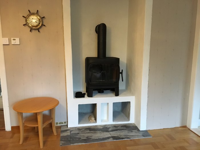 Nyinstallerad svart Jøtul F305-kamin på en vit putsvägg med vedförvaring under och grå klinker framför.