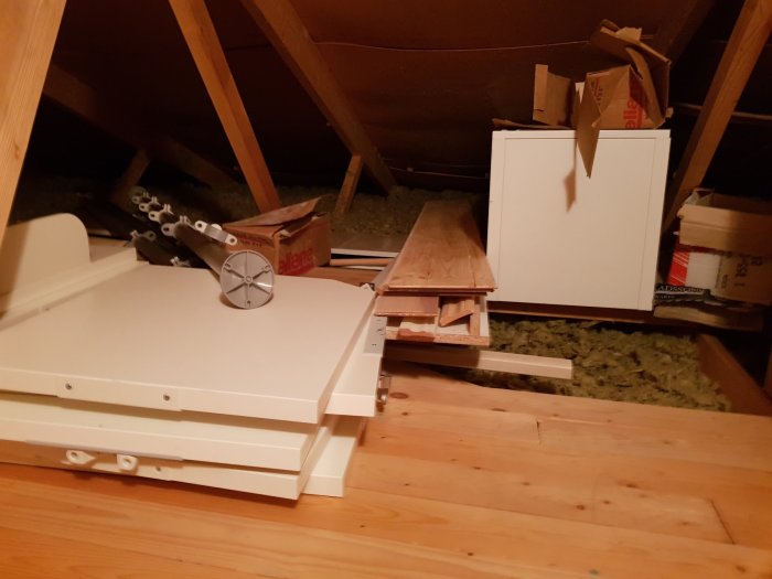 Oorganiserad vind med öppna takbjälkar, isolering och utspridda föremål som kartonger och möbeldelar.