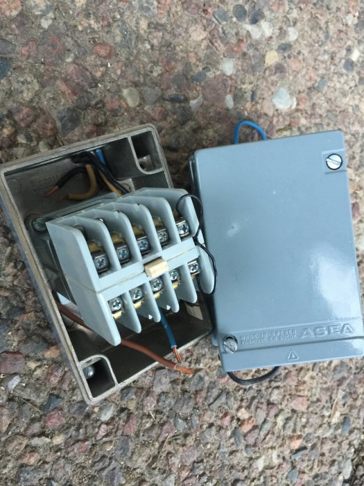 En öppen kontaktor med synliga ledningar och anslutningar, liggande på en grusig yta.