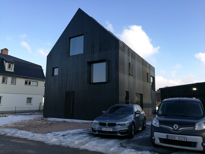 Mörkt modernt hus med skarp arkitektur och fönster, parkerade bilar framför, jämfört med äldre hus.