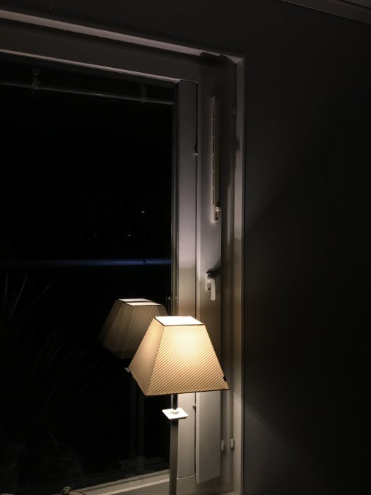 En inomhusbild som visar ett öppet fönster med tilluftsventiler i överkant och en tänd vägglampa.