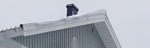 Tak på ett hus med snö och en skorsten mot en grå himmel.