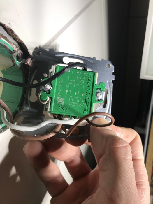 Hand som håller en brun och en vit ledning framför en öppen elektrisk kopplingsdosa med en grön installationsenhet.