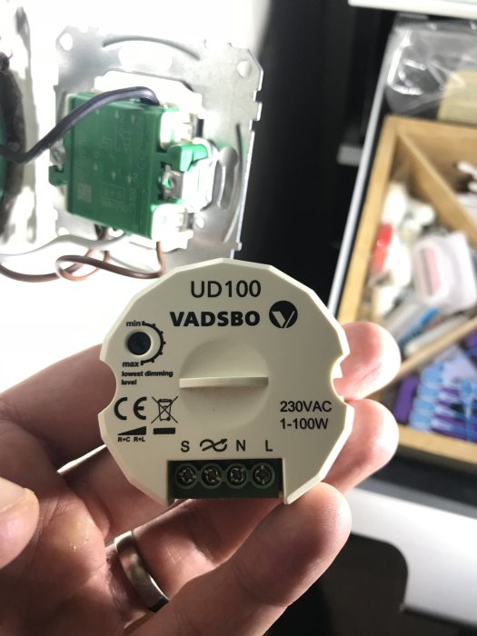 En hand håller en VADSBRO dimmerpuck modell UD100 med synliga anslutningar och etikettering.