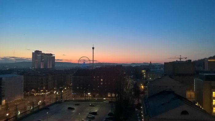 Utsikt över stad vid gryning med byggnader, ett pariserhjul och en färgrik himmel.