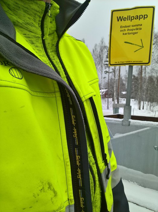 Person i gul reflexjacka nära skylt om wellpappsortering på återvinningsstation.