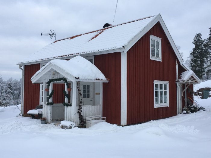 Röd stuga med vita knutar under snötäckt tak i vinterlandskap, diskussion om tillbyggnad vid gaveln.