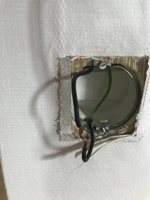 Elinstallation med avskalad kabel runt en skruv i en öppen väggdosas fästram, skadat gips runtomkring.