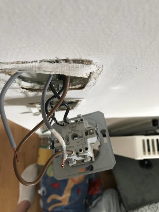 Öppen vägguttag och oskyddade ledningar i olika färger som hänger från ett hål i en gipstak.