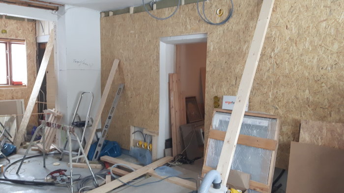 Rörigt arbetsrum under renovering med osb-skivor på väggen, oorganiserade byggmaterial och verktyg.