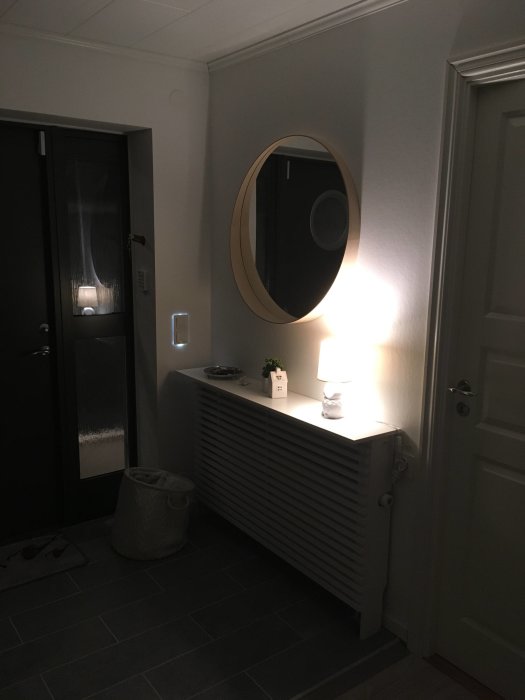 Interiörbild av en hall med rund spegel, radiator, lampa och ytterdörr.