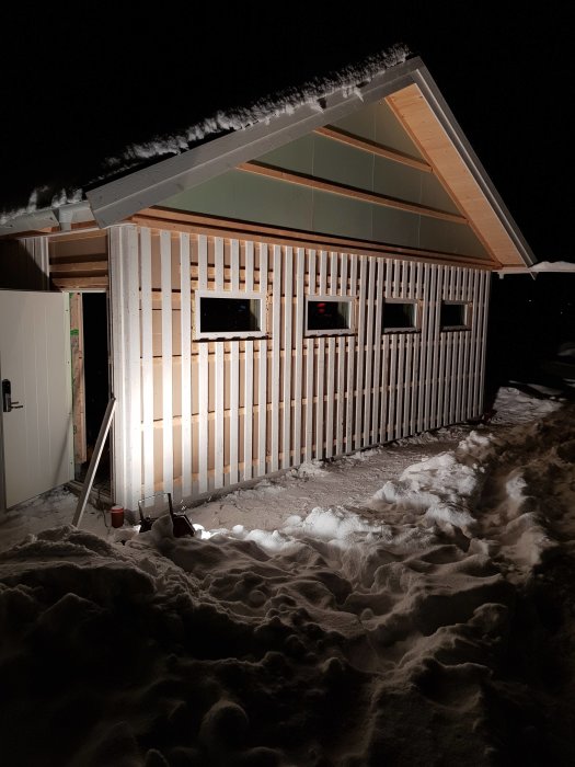 Belyst byggnad på vintern med nylagt underbrädor på gaveln, snötäckt tak och omgivet av snö.