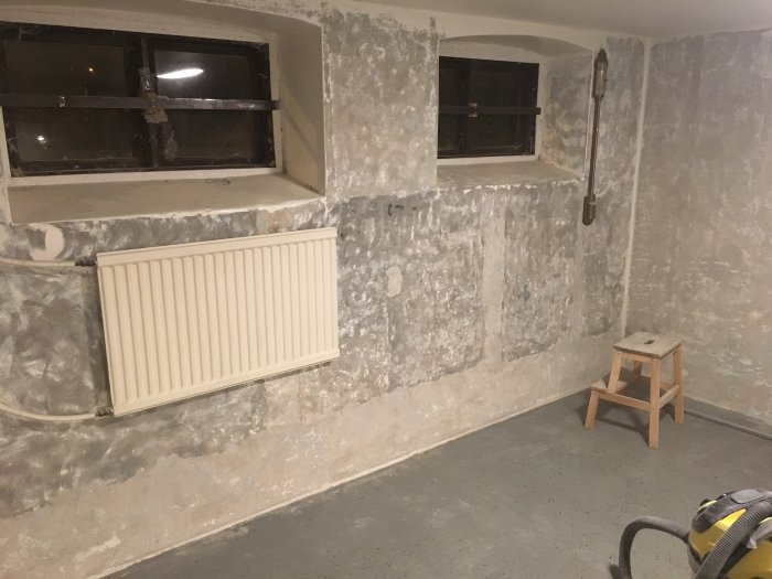 Ett källarrum under renovering med flagnande väggfärg, ett gammalt fönster, radiator och betonggolv.