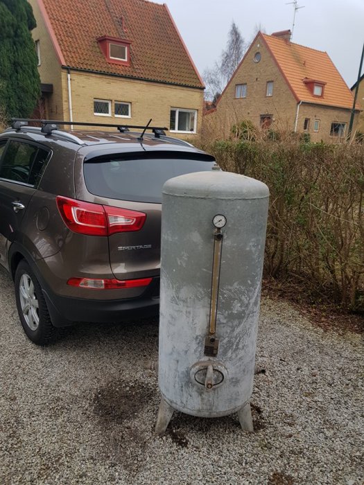 Gammal galvaniserad hydofortank från 1956 står framför en bil på en grusdriveway.