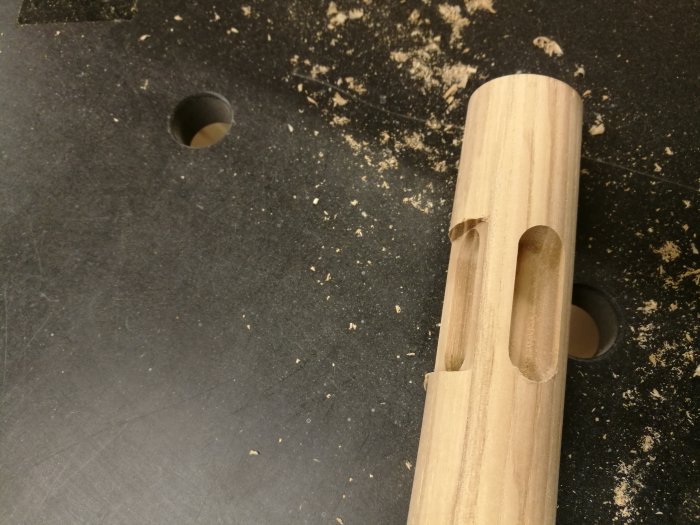 Rundstav i ek med precisionssågade fräsningar och spån på svart arbetsbänk.