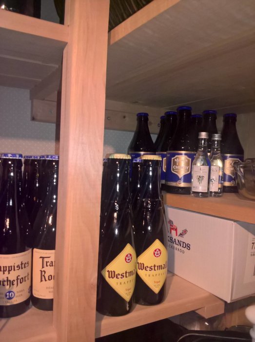 Flaskor av trappistöl på trähylla, inklusive märkena Westmalle och Rochefort, bredvid en kartong med riskakor.