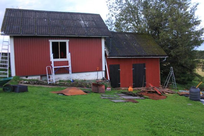 Renoveringsarbete på en röd vedbod, delvis ny panel och stegar, ojämn gräsmatta med byggmaterial.