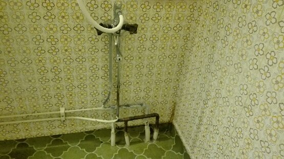 Gammal rörinstallation i hörn av ett våtrum med tapet och klinkergolv, visar sammankopplade rör för varmt och kallt vatten.