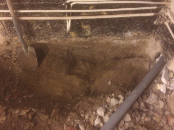 Grävning i byggprojekt med spade, synligt berg och rörledningar i en grop.