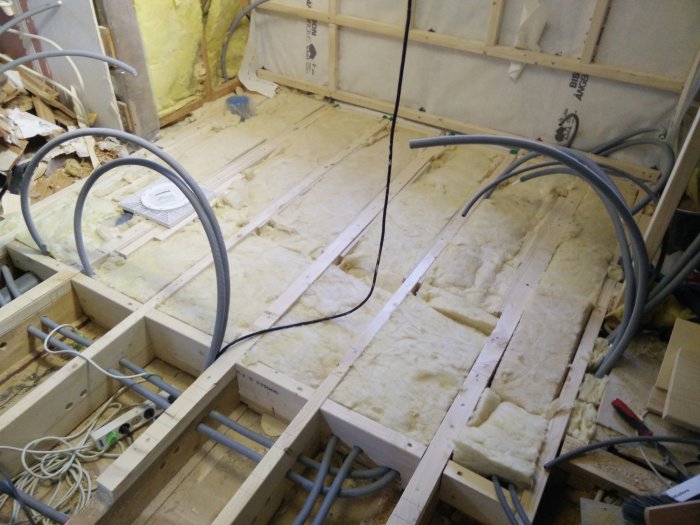 Rakenteilla oleva kylpyhuone, jossa eristeitä on asennettu lattiaan. Puurakenteet ja putket ovat näkyvissä keskeneräisessä tilassa.